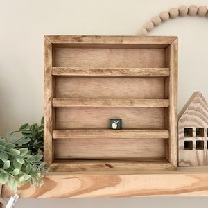May include: A rectangular wooden display shelf with a natural light brown finish, featuring four horizontal tiers. A single small square dark green block with a white graphic of a cupcake is placed on the third shelf from the top.