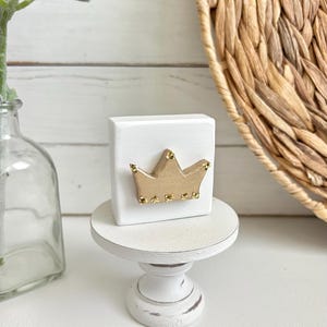 May include: A small white wooden pedestal with a square wooden block on top. The block has a gold crown with rhinestones on it.