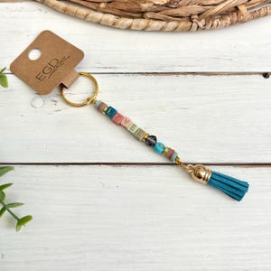 May include: A handmade keychain with the word "LOVED" spelled out in colorful beads. The keychain features a gold-colored ring and a teal tassel. A brown tag with the letters "EGD" is attached to the ring.