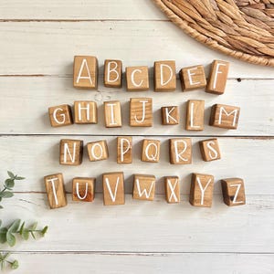 May include: Wooden alphabet blocks with white painted letters on a light wood background. The blocks are arranged in alphabetical order.