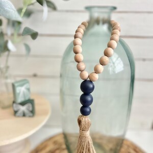 May include: A wooden bead garland with a navy blue tassel. The garland is strung on a clear glass vase.