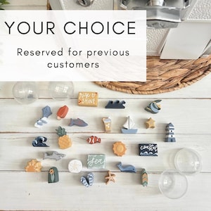 May include: A collection of small, painted wooden figures featuring nautical and beach-themed shapes including a lighthouse, sailboat, fish, pineapple, and flip-flops, alongside two clear plastic capsules and text reading YOUR CHOICE Reserved for previous customers.