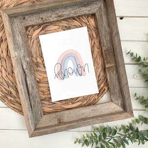 May include: A watercolor rainbow with a pink, peach, and blue color scheme. The rainbow is in a wooden frame with a woven mat. The word "known" is written in black cursive below the rainbow.