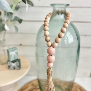 May include: A wooden bead garland with a pink tassel. The garland is made of natural wood beads and has a pink tassel at the end. The garland is hanging from a clear glass vase.
