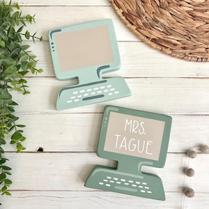 May include: Two sage green wooden computer-shaped ornaments. One features a blank beige screen, while the other displays the white text "MRS. TAGUE" on a beige screen. Both items have a stylized keyboard base with white rectangular keys.