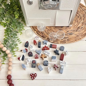 May include: A collection of small, hand-painted wooden shapes in red, white, and blue, featuring stars, hearts, flowers, and patriotic symbols. One piece is rectangular with the word AMERICA printed in black. These items are scattered on a white surface.