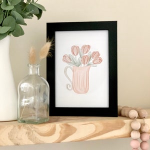 May include: A framed watercolor print of a pink pitcher filled with pink tulips. The pitcher has a textured surface and a curved handle. The print is on a white background and is framed in black.