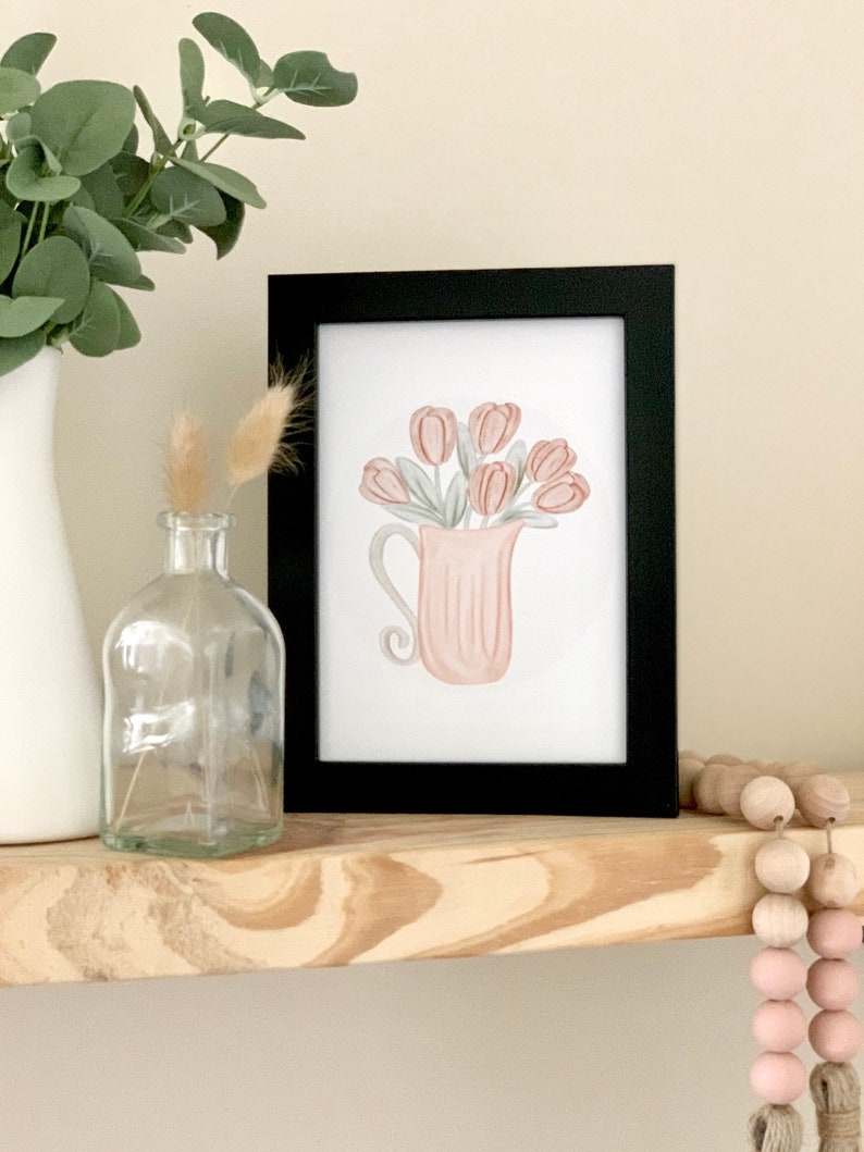 May include: A framed watercolor print of a pink pitcher filled with pink tulips. The pitcher has a textured surface and a curved handle. The print is on a white background and is framed in black.