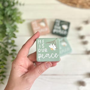 May include: A square sage green wooden block featuring the white text "HE IS OUR PEACE" and a small painted white flower with green leaves. The block is held in a hand, with other similar colored blocks visible in the blurred background.