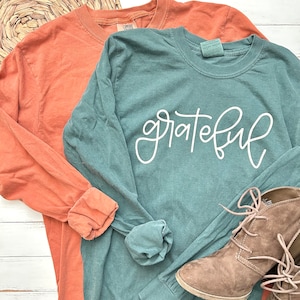 May include: Two long-sleeved shirts, one in a muted orange and the other in teal, are displayed. The teal shirt features the word "grateful" in white script. A pair of brown suede boots with laces is visible in the bottom right corner.