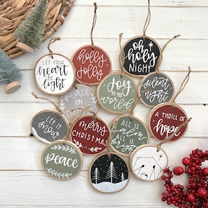 Peut inclure: Une collection de décorations de Noël rondes en bois, avec diverses phrases de vacances en écriture blanche. Les ornements sont peints dans des tons de rouge, vert, gris et noir, et sont suspendus avec de la ficelle. Phrases incluent "Paix" et "Joyeux Noël".
