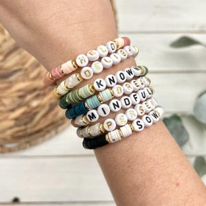 Peut inclure: Un ensemble de bracelets en perles colorées avec des perles de lettres blanches qui épellent les mots "Held Delight", "Know", "Dream", "Mindful", "Present", et "Soul".