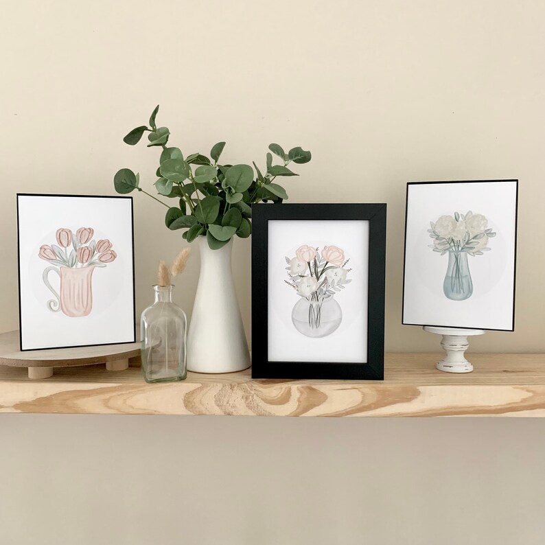 May include: Three framed prints of watercolor floral arrangements in different colored vases. The prints are displayed on a wooden shelf with a vase and a bottle.