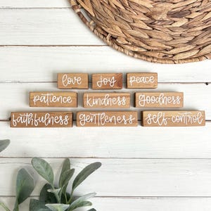 May include: Wooden blocks with white cursive text spelling out words like "love", "joy", "peace", "patience", "kindness", "goodness", "faithfulness", "gentleness", and "self-control". The blocks are arranged on a white wooden surface.