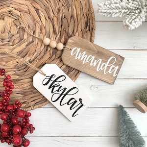 May include: Two wooden gift tags, one stained brown and one white, with the names "Amanda" and "Skylar" in elegant white and black script. Each tag has a jute string and wooden bead detail. The tags rest on a woven placemat.
