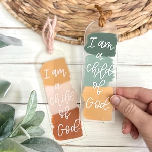 May include: Two clear acrylic bookmarks with the text "I am a child of God." One bookmark has peach, pink, and brown brushstrokes, and the other has green, blue, and yellow brushstrokes. Each bookmark has a decorative string.