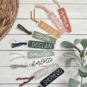 May include: Ten clear acrylic bookmarks with colorful painted stripes and white handwritten names. The names are: Piper, tyla, Declan, emily, charlotte, georgia, SHEPHERD, olivia, mama.