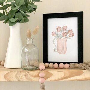 May include: A framed watercolor print of a pink pitcher filled with pink tulips. The print is on a wooden shelf with a white vase, a clear glass bottle, and a pink and natural wood bead garland with a tassel.