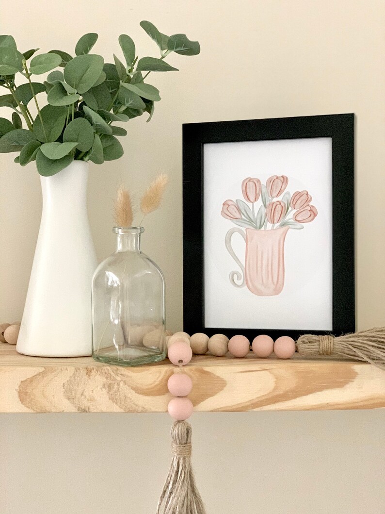 May include: A framed watercolor print of a pink pitcher filled with pink tulips. The print is on a wooden shelf with a white vase, a clear glass bottle, and a pink and natural wood bead garland with a tassel.
