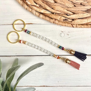 May include: Two keychains with gold rings, clear beaded strands with white letter beads, and colorful accents. One keychain has a navy blue tassel, the other a peach tassel. The background includes a woven basket and white wood.