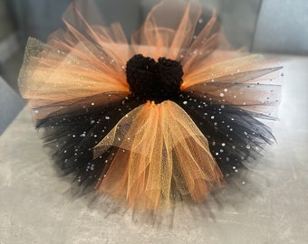 May include: A black and orange tulle tutu with a black felt heart in the centre. The tutu is decorated with iridescent sequins.