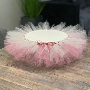 May include: A white cake stand with a pink tulle skirt and a pink satin bow. The stand is on a wooden table.