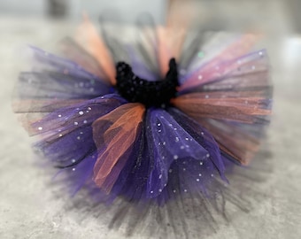Puede incluir: Un tutú de varias capas en tonos morado, negro y naranja, con un adorno negro en forma de media luna. El tul está salpicado de purpurina plateada. Este accesorio de disfraz es adecuado para un niño.