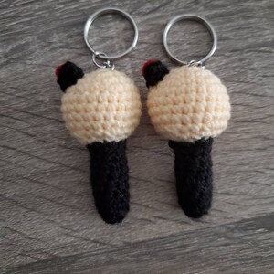 May include: Two handmade crochet keychains. Each keychain features a cream-colored, round head with black ears and a red accent, atop a black cylindrical body. Silver key rings are attached to the top. The keychains are displayed on a wooden surface.