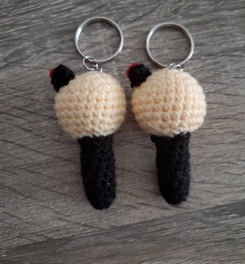 May include: Two handmade crochet keychains. Each keychain features a cream-colored, round head with black ears and a red accent, atop a black cylindrical body. Silver key rings are attached to the top. The keychains are displayed on a wooden surface.
