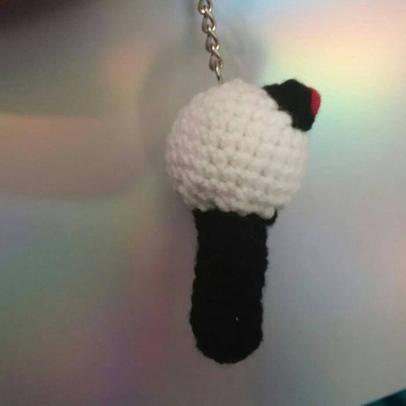 May include: Crocheted keychain featuring a white sphere with a black base and a small red accent. The keychain is handmade and attached to a silver chain. The design is a stylized representation of a microphone.