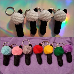 May include: A collection of crochet microphone keychains in various colors. The microphones have a white or colored ball-shaped top, a black handle, and a silver keyring. The keychains are displayed on a reflective surface and a purple fabric.