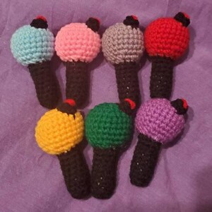 May include: A collection of seven handmade crochet microphones in various colors, including blue, pink, gray, red, yellow, green, and purple. Each microphone has a black handle and a small red and black accent on top.