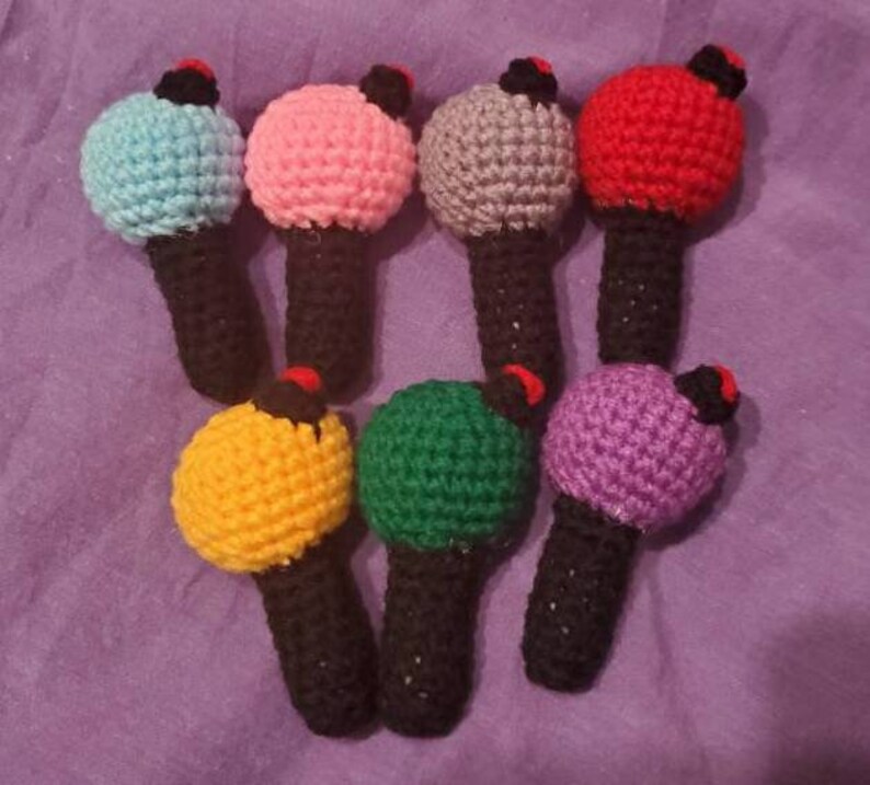 May include: A collection of seven handmade crochet microphones in various colors, including blue, pink, gray, red, yellow, green, and purple. Each microphone has a black handle and a small red and black accent on top.