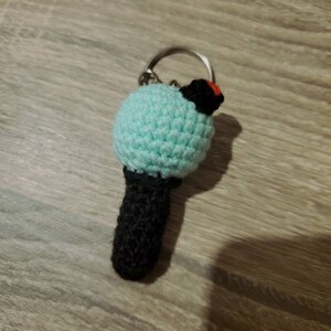 May include: A handmade crochet keychain featuring a light blue sphere, a black cylindrical base, and a small black and red accent. The keychain is attached to a silver metal ring. The item is crafted with a visible stitch pattern.