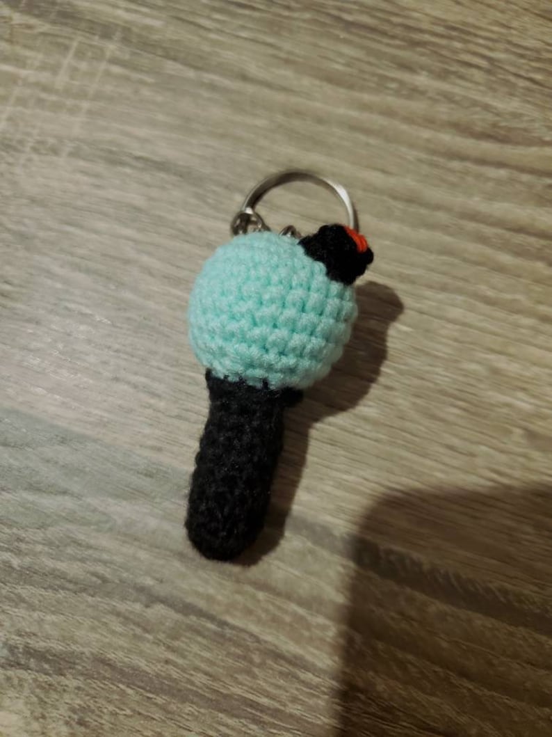 May include: A handmade crochet keychain featuring a light blue sphere, a black cylindrical base, and a small black and red accent. The keychain is attached to a silver metal ring. The item is crafted with a visible stitch pattern.