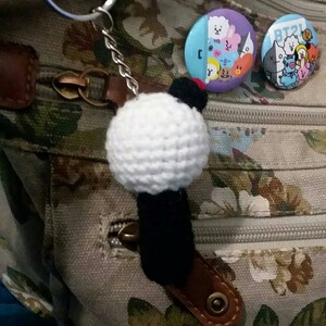 May include: A handmade white crochet ball keychain with a black base and a small red accent. Two round BT21 character pins are also visible. The keychain is attached to a brown leather strap.