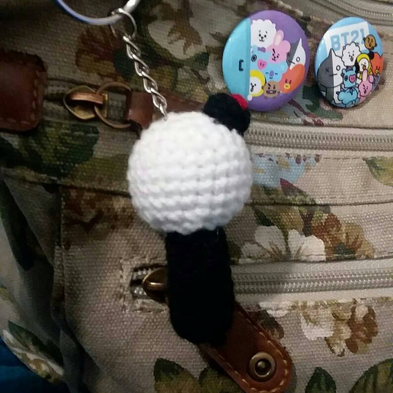 May include: A handmade white crochet ball keychain with a black base and a small red accent. Two round BT21 character pins are also visible. The keychain is attached to a brown leather strap.