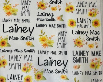 May include: A white blanket with a sunflower pattern and the name "Lainey Mae Smith" repeated throughout. The name is written in black text.