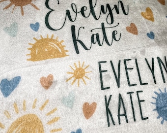 May include: A white fabric blanket with a repeating pattern of suns, hearts, and the names "Evelyn Kate" written in black cursive.
