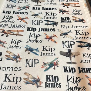 May include: A white blanket featuring a repeating pattern of vintage airplanes in various colors, including blue, orange, and brown. The name "Kip James" is printed in black script throughout the design. The blanket is a soft, cozy textile.