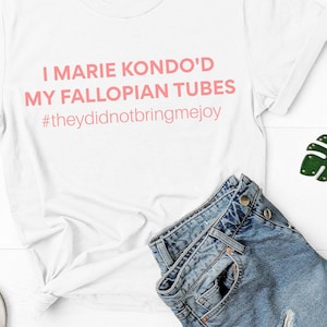 May include: White t-shirt with the text "I Marie Kondo'd My Fallopian Tubes #theydidnotbringmejoy" in pink. The image also includes denim shorts, silver sandals, red sunglasses, and a green leaf keychain.