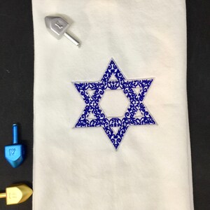 May include: White fabric with a blue embroidered Star of David with a decorative pattern. The Star of David is centered on the fabric.