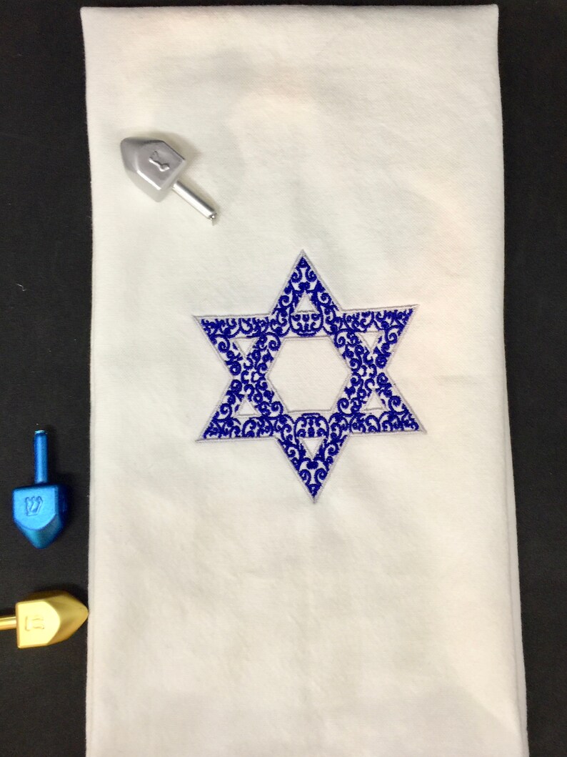 May include: White fabric with a blue embroidered Star of David with a decorative pattern. The Star of David is centered on the fabric.