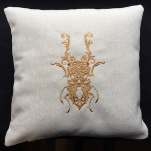 May include: A white throw pillow with a gold embroidered design of a stylized insect with red accents. The pillow is on a black background.