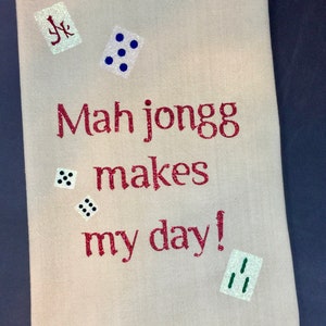 May include: A beige kitchen towel with the text "Mah jongg makes my day!" in red glitter. The towel is decorated with various dice in blue, black, and green glitter.