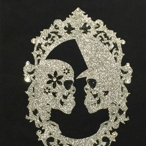 May include: Black fabric with a silver glitter design featuring two skulls facing each other within an ornate oval frame. One skull has a flower and the other wears a hat. Rhinestones accent the frame.