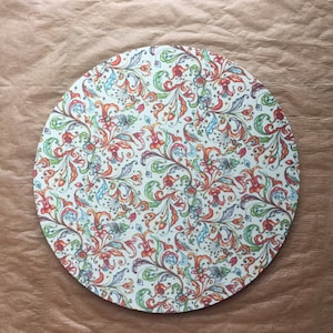 May include: Round white placemat with a colorful floral pattern. The pattern features red, orange, yellow, green, blue, and purple flowers and leaves.