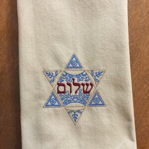 May include: A white kitchen towel with a blue and gold embroidered Star of David. The Hebrew word "Shalom" is embroidered in red inside the star.