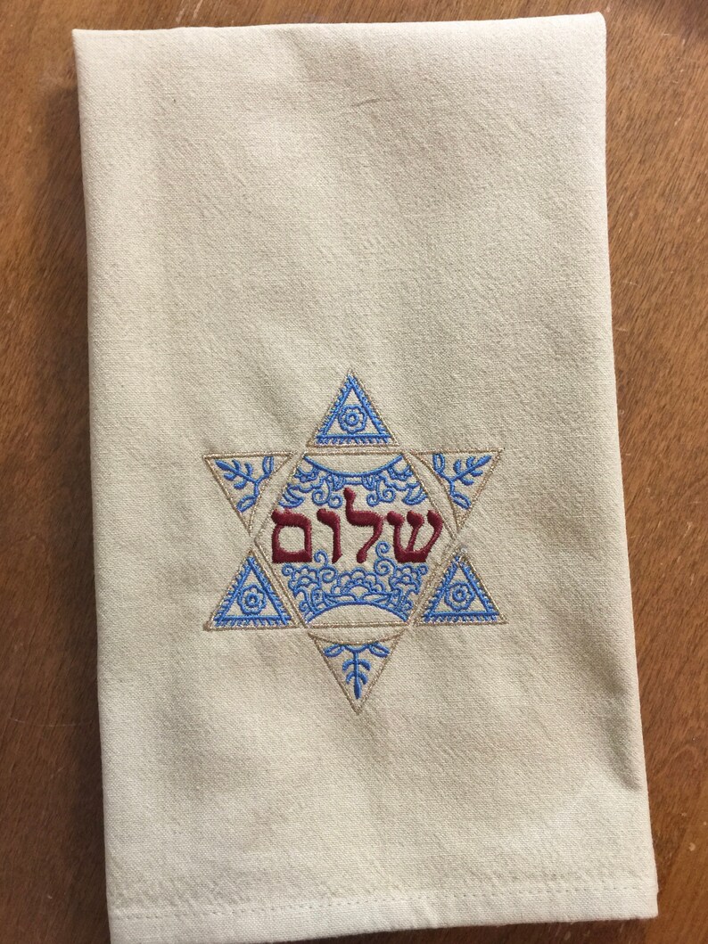 May include: A white kitchen towel with a blue and gold embroidered Star of David. The Hebrew word "Shalom" is embroidered in red inside the star.