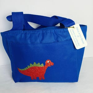 May include: Blue insulated lunch bag with a red glitter dinosaur applique. Features a front pocket and a blue handle. A tag reads "Insulated Bags, Choice of 6 Colors, Personalization Included."